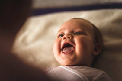 High angle view of cheerful newborn baby girl lying down on the bed and laughing while her mother, that is blurred in foreground, is playing with her.