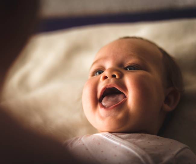 High angle view of cheerful newborn baby girl lying down on the bed and laughing while her mother, that is blurred in foreground, is playing with her.