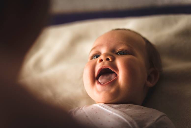 High angle view of cheerful newborn baby girl lying down on the bed and laughing while her mother, that is blurred in foreground, is playing with her.