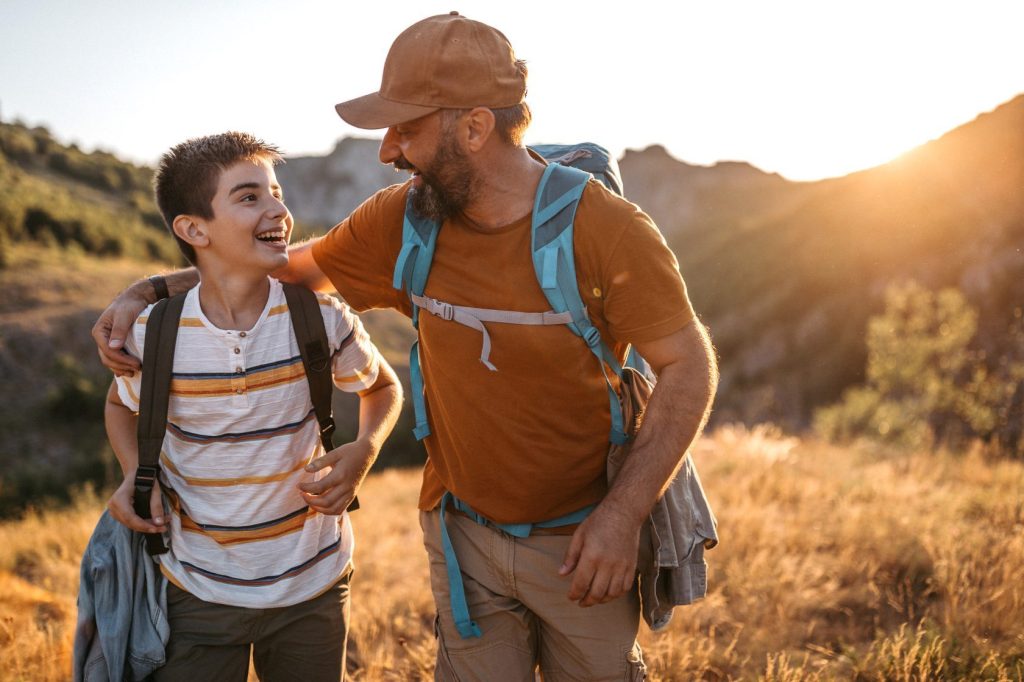 Ragazzo adolescente allegro che cammina nella natura con suo padre, abbracciandolo, parlando e sorridendo